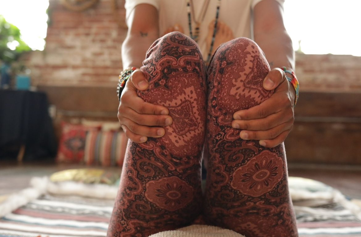 Ryan holding his knees, seated on a woven rug in a brick-walled studio